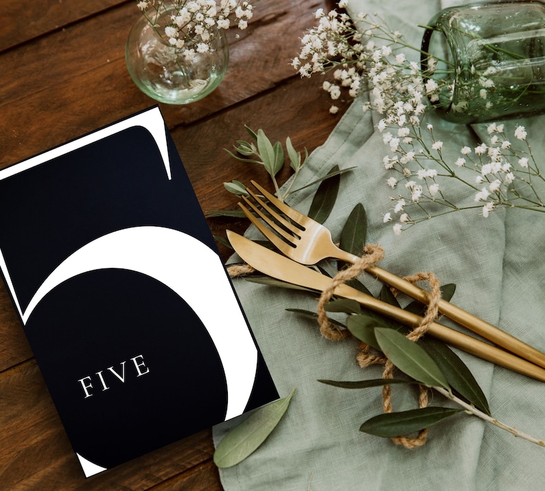 May include: A black and white table number card with the number "5" and the word "FIVE" in white text. The card is on a wooden table with a green cloth, gold cutlery, and white flowers.