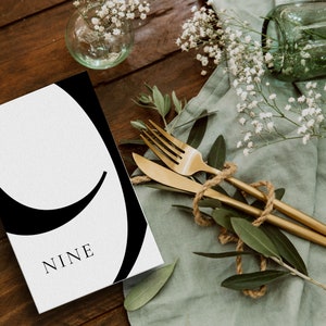 Puede incluir: Una tarjeta blanca con un gr&aacute;fico negro del n&uacute;mero nueve. La tarjeta est&aacute; sobre una mesa de madera con un pa&ntilde;o verde y vegetaci&oacute;n.