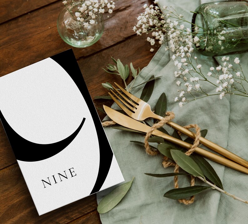Puede incluir: Una tarjeta blanca con un gr&aacute;fico negro del n&uacute;mero nueve. La tarjeta est&aacute; sobre una mesa de madera con un pa&ntilde;o verde y vegetaci&oacute;n.