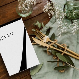 Puede incluir: Una tarjeta blanca con texto negro que dice "SEVEN" y una l&iacute;nea diagonal negra. La tarjeta est&aacute; sobre una mesa de madera con una servilleta de tela verde, cubiertos de oro y hojas verdes.