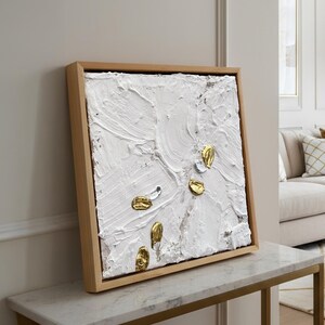 Könnte beinhalten: Quadratisches abstraktes Kunstwerk mit strukturierter weißer Oberfläche und goldenen und silbernen Akzenten, gerahmt in Naturholz. Das Kunstwerk wird auf einem Tisch mit Marmorplatte präsentiert und zeigt eine moderne Ästhetik.