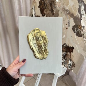 May include: A small, rectangular canvas with a light gray background. A textured, gold-colored abstract shape is centered on the canvas. The artwork is held in a person's hand, with a second painting visible in the background.