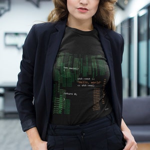 Peut inclure: Une femme portant un blazer noir et un pantalon noir. Elle porte un t-shirt noir avec du code vert imprimé dessus. Le code comprend le texte "Hello, world".