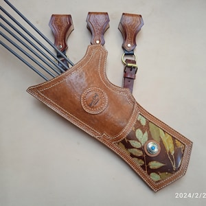 May include: A tan leather archery quiver featuring a circular embossed logo, white stitching, and a lower panel with a printed leaf pattern accented by a small silver-toned concho with a turquoise center. Five dark grey arrows are inserted inside.