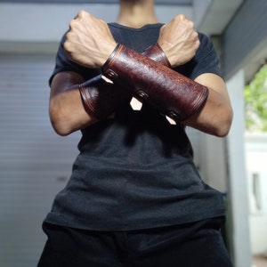 May include: A pair of brown leather arm guards with decorative stitching and buckles. The arm guards are worn on both arms and are crossed over each other.
