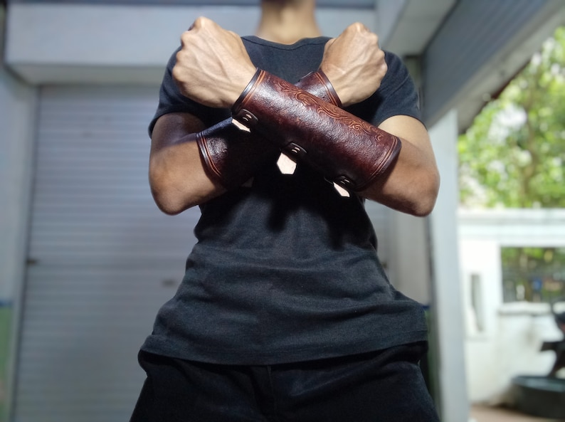 May include: A pair of brown leather arm guards with decorative stitching and buckles. The arm guards are worn on both arms and are crossed over each other.