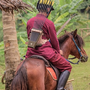 May include: A person wearing a red shirt and black boots rides a brown horse. The person is holding a bow and arrow, and the arrows are strapped to their back. The horse is standing in a field of green grass.