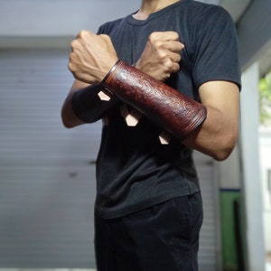 May include: A pair of brown leather arm guards with intricate designs. The guards are worn on both arms and are secured with straps.