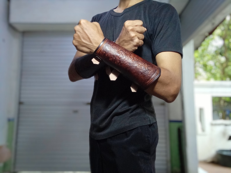 May include: A pair of brown leather arm guards with intricate designs. The guards are worn on both arms and are secured with straps.