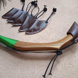 May include: A wooden recurve bow with a green grip and leather arrow rests. The bow has a brown finish and is paired with several brown leather arrow rests with white stitching and black cords.