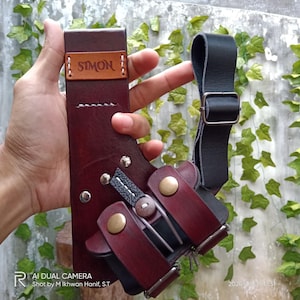 Könnte beinhalten: Ein braunes Lederholster mit einem schwarzen Riemen und einer Metallschnalle. Das Holster hat eine Tasche auf der Vorderseite mit einem Knopfverschluss. Das Holster ist mit "SIMON" beschriftet.