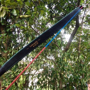 May include: A black bow with a green edge and the text "Kalimasada ~ 45.28" is held against a backdrop of green foliage. A red and blue string extends from the bow.