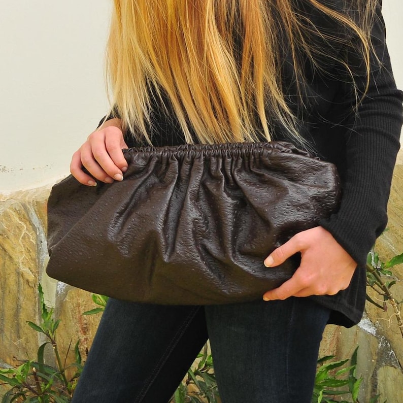 May include: A dark brown, textured clutch bag with a gathered top. The bag is held by a person wearing a black long-sleeved shirt and dark jeans. The bag has a soft, rounded shape.