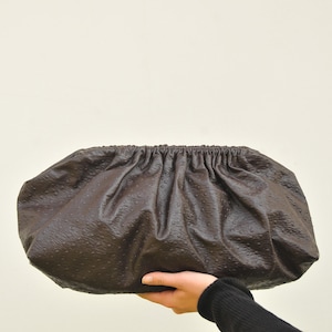 May include: A dark brown clutch purse with a textured surface. The purse has a gathered top and is held by a person's hand. The bag is oval-shaped and appears to be made of leather.