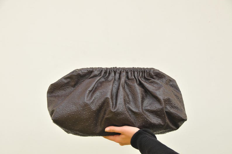 May include: A dark brown clutch purse with a textured surface. The purse has a gathered top and is held by a person's hand. The bag is oval-shaped and appears to be made of leather.