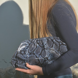 Può includere: Una pochette con motivo a pelle di serpente blu e grigio. La borsa è tenuta davanti a una persona che indossa una maglia a maniche lunghe blu navy. La borsa ha una parte superiore arricciata e una forma morbida e arrotondata.
