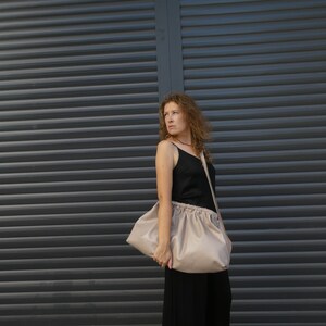 May include: A woman wearing a black tank top and black pants is holding a large, light beige, slouchy bag with a long strap.