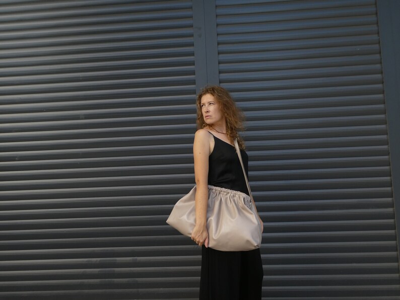 May include: A woman wearing a black tank top and black pants is holding a large, light beige, slouchy bag with a long strap.