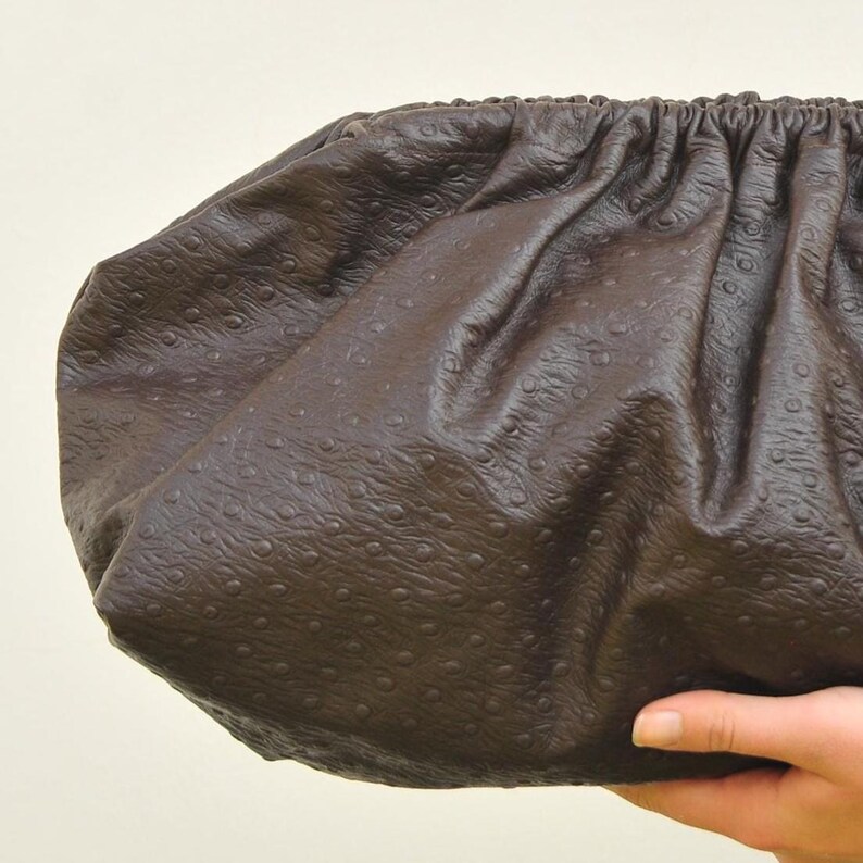 May include: A dark brown clutch bag with a textured, ostrich-like pattern. The bag has a gathered top and a rounded shape. The material appears to be leather or a leather-like material.