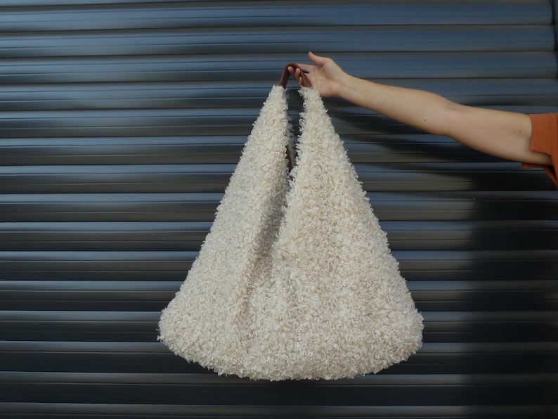 May include: A large, fluffy, off-white tote bag with brown leather handles. The bag is made of a soft, fuzzy material and has a relaxed, casual look.
