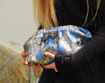 May include: A clutch bag with a snakeskin pattern in shades of blue, gray, and white. The bag is gathered at the top and held in the hands. The background is blurred, with a glimpse of a stone wall.