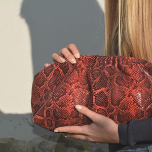 Puede incluir: Un bolso de mano con estampado de piel de serpiente roja y negra. El bolso lo sostiene una persona. El bolso tiene forma redondeada y una superficie texturizada. La combinación de colores es principalmente roja y negra, con un acabado brillante.