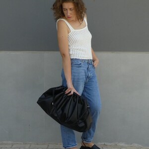 May include: A woman wearing a white crochet tank top, blue jeans, and black sandals is holding a large black bag.