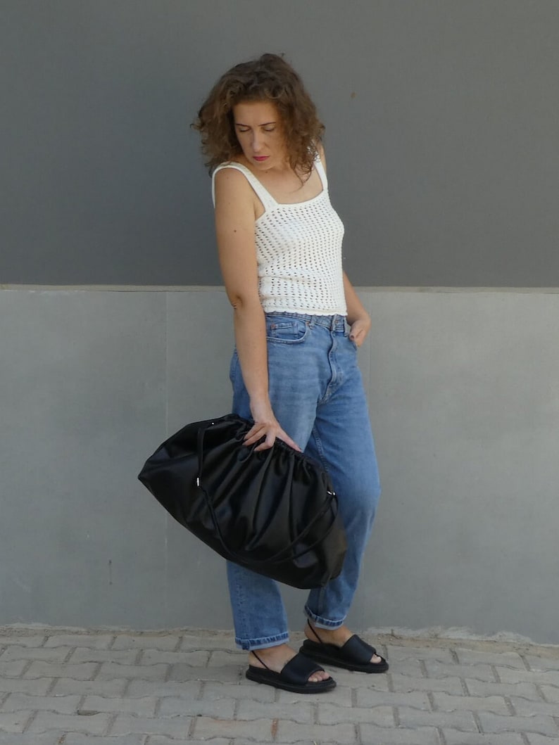 May include: A woman wearing a white crochet tank top, blue jeans, and black sandals is holding a large black bag.