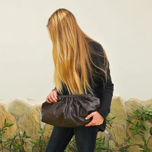 May include: A dark brown, gathered leather clutch bag. The bag is held by a person wearing a black long-sleeve shirt and dark blue jeans. The background is a light-colored wall and stone.