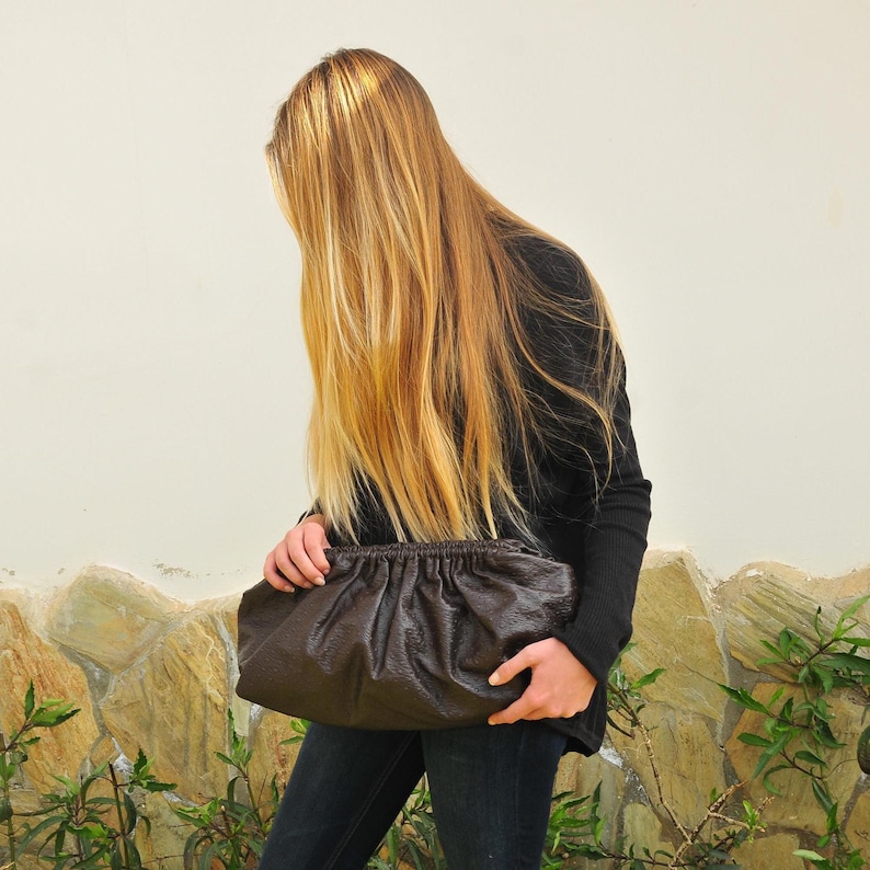 May include: A dark brown, gathered leather clutch bag. The bag is held by a person wearing a black long-sleeve shirt and dark blue jeans. The background is a light-colored wall and stone.