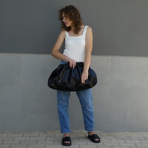 May include: A woman is holding a black, gathered clutch purse. She is wearing a white crocheted tank top and blue jeans.