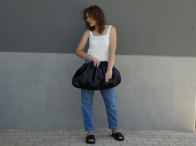 May include: A woman is holding a black, gathered clutch purse. She is wearing a white crocheted tank top and blue jeans.