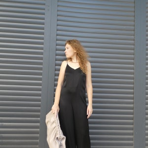 May include: A woman wearing a black sleeveless jumpsuit stands in front of a gray metal roller shutter door. She is holding a light brown bag in her left hand.