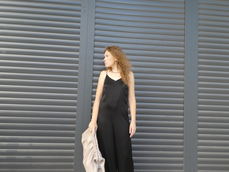 May include: A woman wearing a black sleeveless jumpsuit stands in front of a gray metal roller shutter door. She is holding a light brown bag in her left hand.