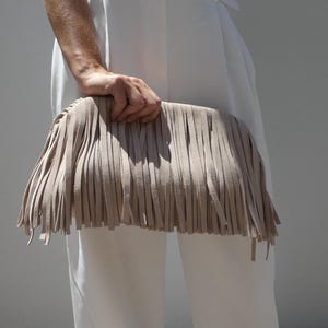 Peut inclure: Une pochette beige à franges. Le sac est rectangulaire avec de longues franges verticales. La frange est d'une nuance de beige plus claire que le sac lui-même. Le sac est tenu devant une personne vêtue de blanc.