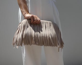 May include: A beige fringed clutch bag. The bag is rectangular with long, vertical fringe. The fringe is a lighter shade of beige than the bag itself. The bag is held in front of a person wearing white clothing.