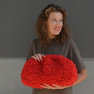 Puede incluir: Una mujer sostiene un bolso de mano rojo brillante y esponjoso. El bolso está hecho de un material suave y peludo y tiene una forma redondeada.