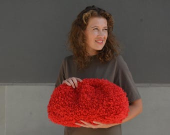 May include: A bright red, textured, fuzzy clutch bag with a rounded, voluminous shape. The exterior features a dense, curly pile material that creates a soft, tactile appearance. The bag is held horizontally, showcasing its curved, pouch-like design.