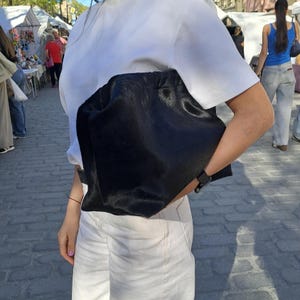Puede incluir: Un bolso de mano grande, negro y de lados blandos con un acabado texturizado similar al pelaje. El bolso presenta una abertura fruncida estilo bolsa en la parte superior y se sostiene horizontalmente contra el costado del cuerpo, mostrando su color negro sólido.