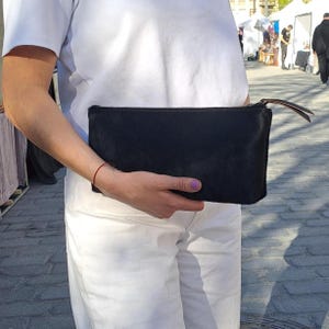 May include: A rectangular black clutch bag with a textured hair-on-hide finish. The bag features a slim profile, a top zip closure with a thin leather pull tab, and a clean, minimalist design. The surface has a soft, matte appearance.