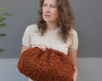 May include: A rust-colored, textured, curly-fleece clutch bag with a rounded, soft-sided shape. The bag features a dense, shaggy exterior surface and is held in front of a plain, light-colored, short-sleeved knit top.