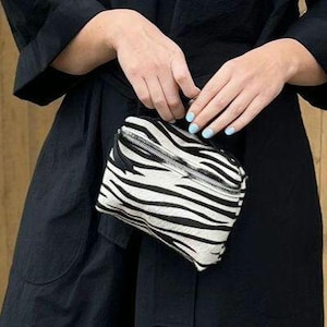 Puede incluir: Un pequeño bolso rectangular con un patrón de rayas de cebra en blanco y negro. El bolso está confeccionado con un material texturizado e incluye un cierre de cremallera de metal plateado colocado en diagonal en la parte frontal del accesorio.