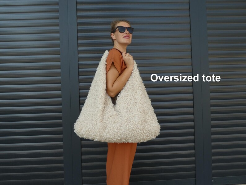 May include: A white fluffy oversized tote bag. The bag is made of a soft, fuzzy material and has a large, open top. The bag is being held by a person wearing a brown shirt and sunglasses.