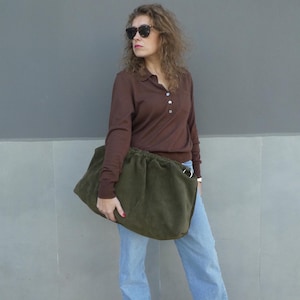 May include: A woman wearing a brown long-sleeved shirt and blue jeans is carrying a large, dark green, slouchy bag.