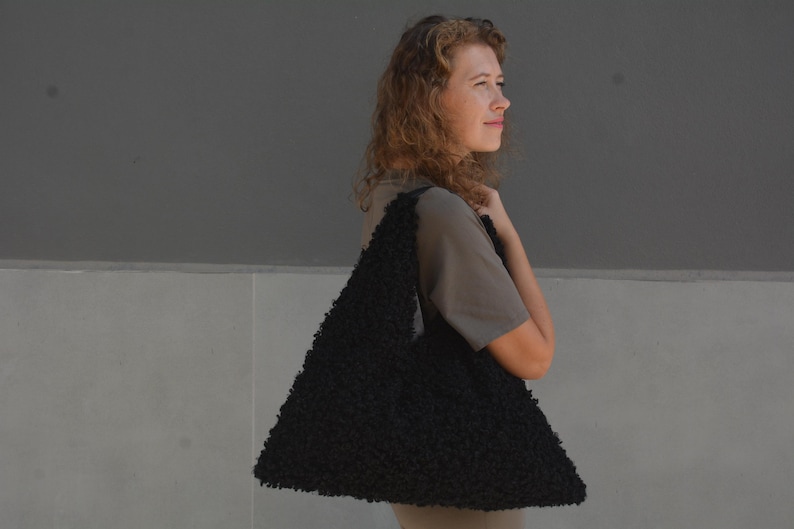 May include: A woman wearing a gray shirt is holding a large black fuzzy tote bag.