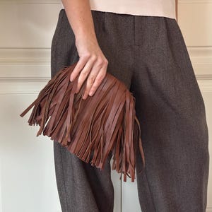 Pode incluir: Uma bolsa clutch castanha com uma camada densa de franjas verticais compridas que cobrem toda a superfície frontal. O acessório é feito de um material liso de cor sólida e mantém uma forma retangular com extremidades arredondadas.