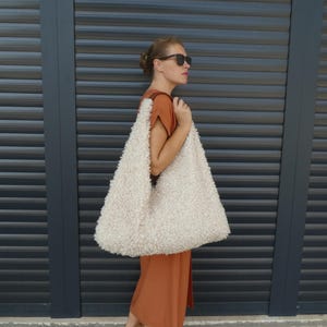 May include: A woman wearing a brown dress and sunglasses is holding a large, white, fluffy tote bag.