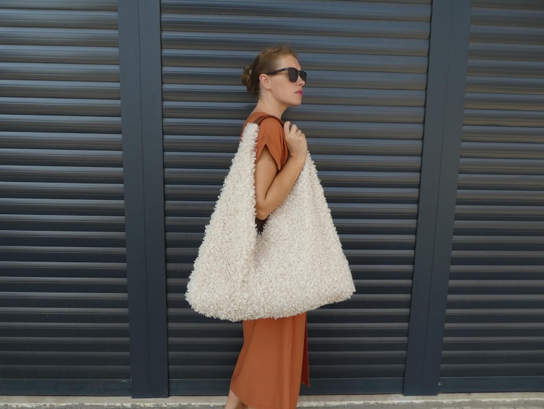 May include: A woman wearing a brown dress and sunglasses is holding a large, white, fluffy tote bag.