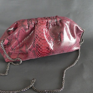 Könnte beinhalten: Eine rote Schlangenleder-Clutch mit einer Kettenkette. Die Tasche hat einen gerafften Oberteil und eine glänzende Oberfläche.