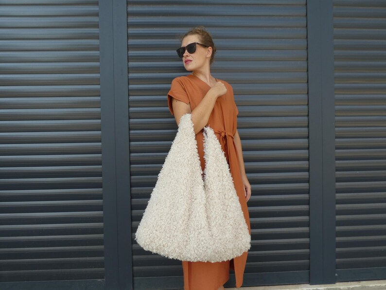 May include: A woman wearing a burnt orange dress and sunglasses holds a large, white, fluffy tote bag.